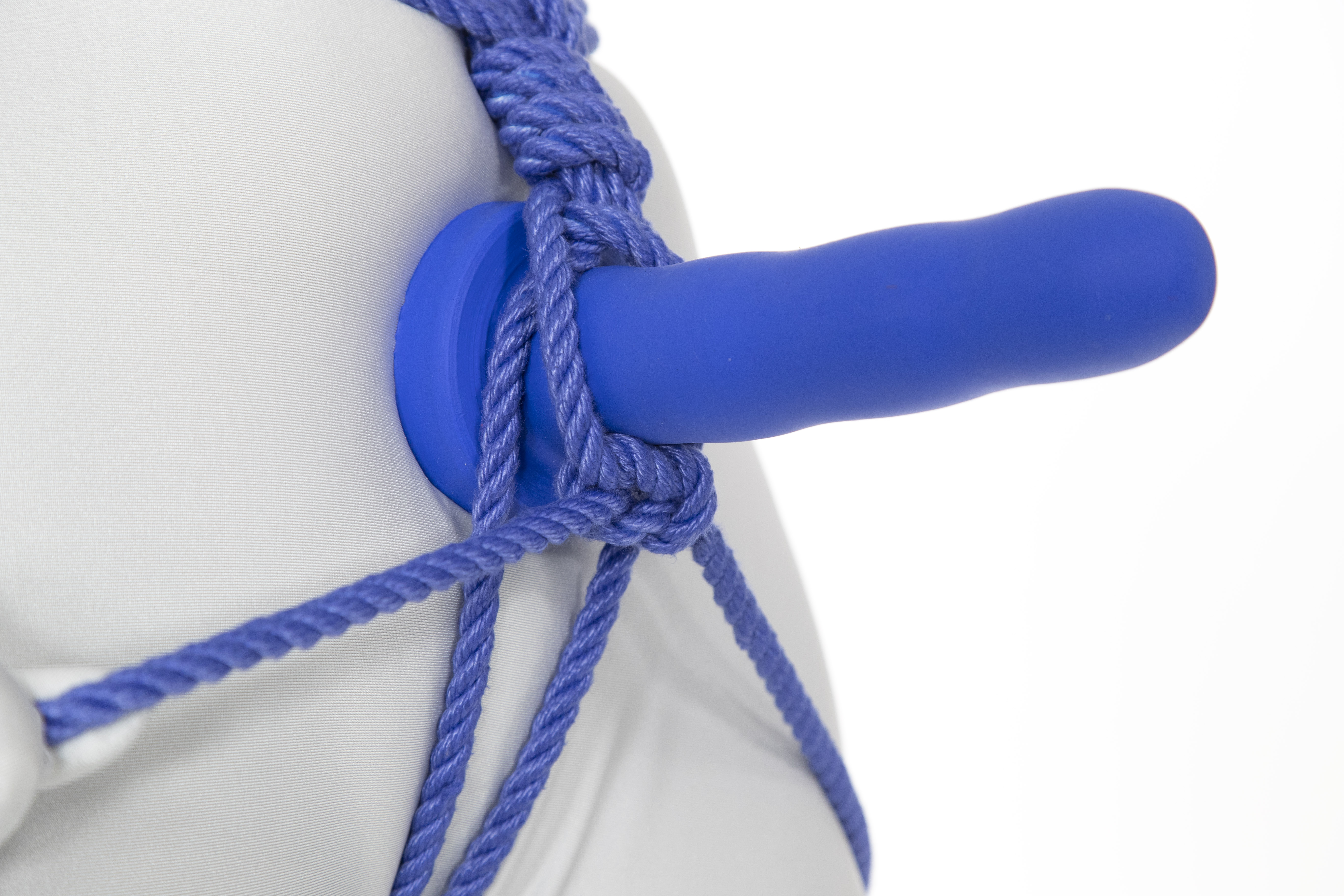 A closeup view of the crotch of a person in a white bodysuit. A blue dildo is mounted over their pubic mound, sticking straight out. A spiral of blue rope runs into the dildo from above. The ends of the rope separate and go down around either side of the dildo, holding it in place with a square knot underneath. The ends of the rope coming out of the square knot separate and pass around either side of the body. Another doubled rope comes up between the legs, separates around the dildo, and travels through the center of the rope spiral.
