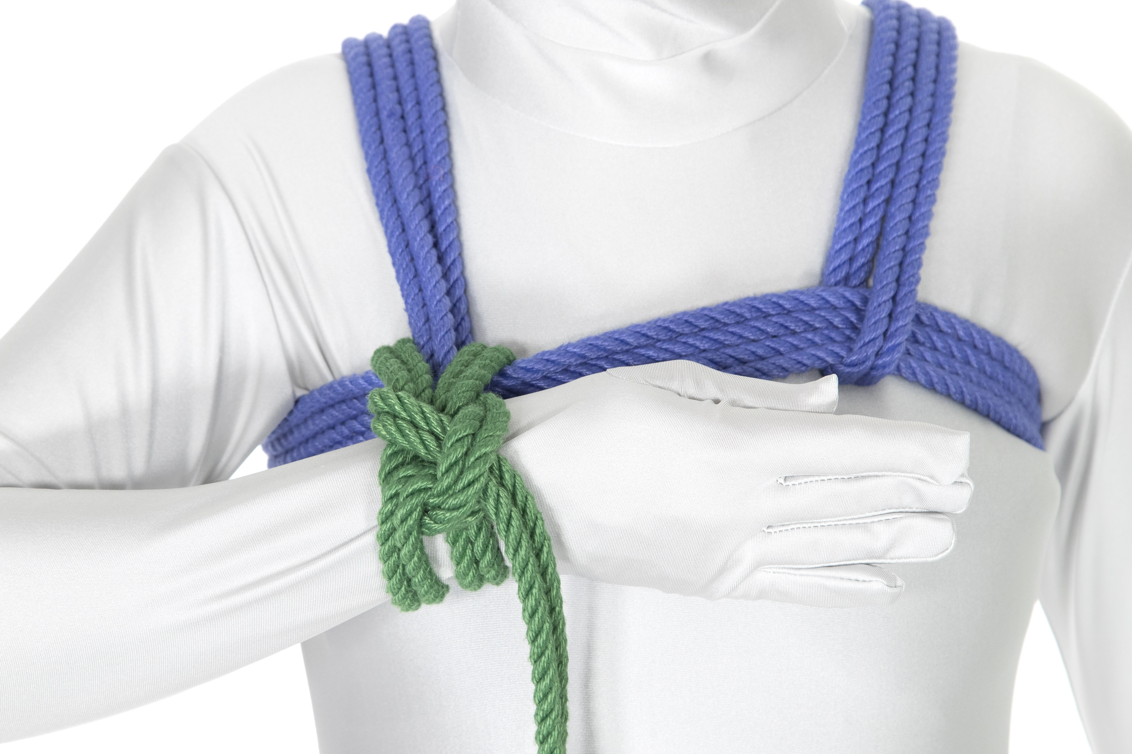 A front view of a person in a white bodysuit wearing a blue chest harness. Their right arm is bent at the elbow. A green column tie binds the right wrist to the chest harness at the point where the right shoulder line connects to the front chest wraps.