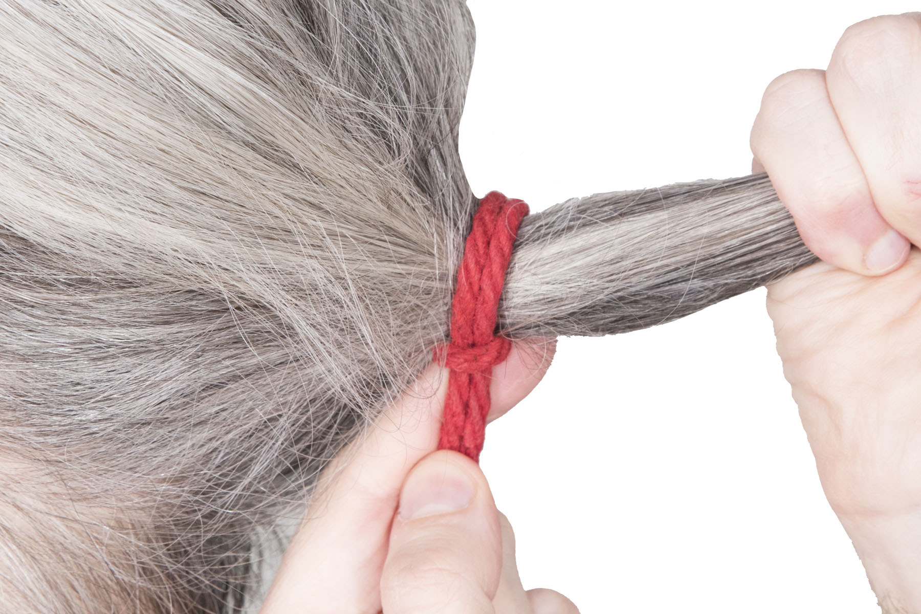 A closeup of a head with long graying hair. The rigger’s right hand is pulling the hair back into a ponytail. The rigger’s left hand has slipped the lark’s head off their wrist and slid it onto the ponytail, right up to the scalp.