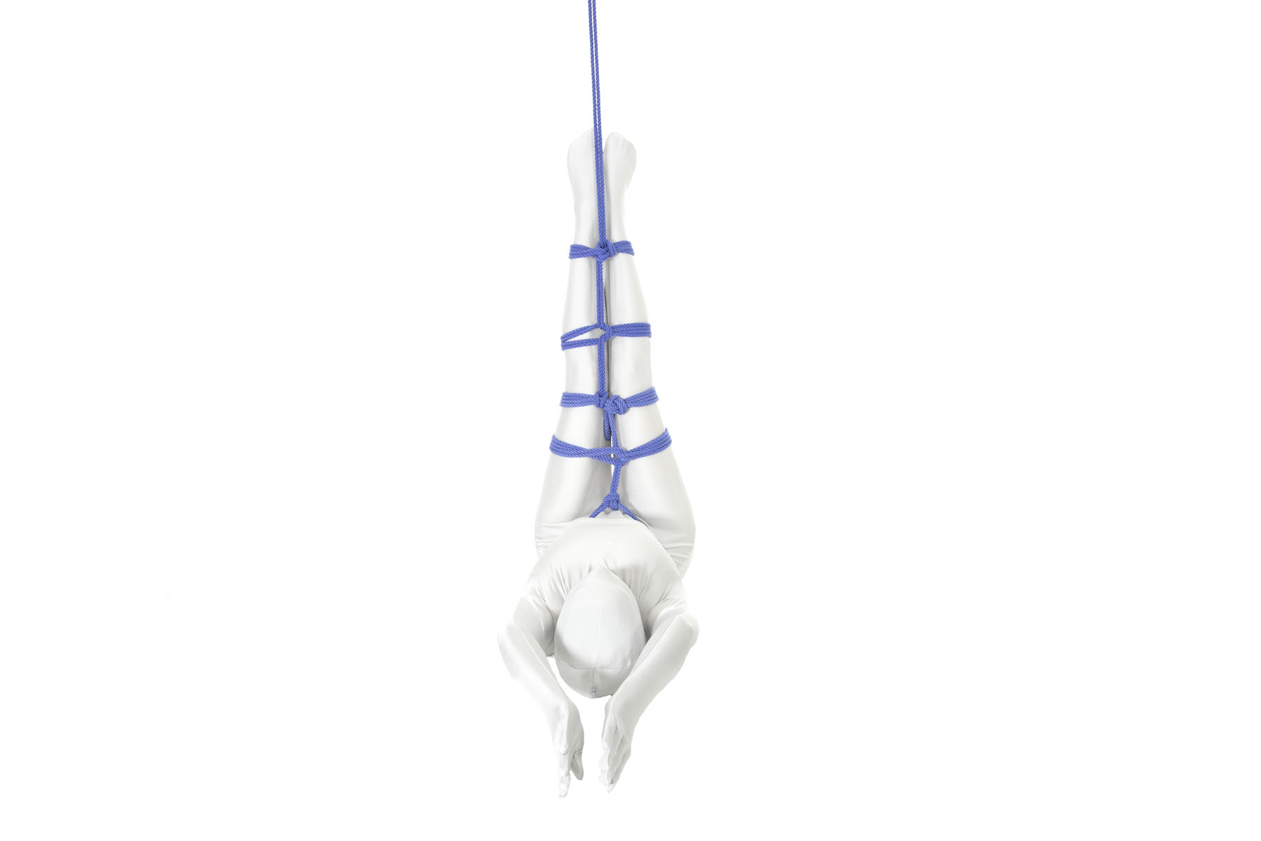 A person in a white bodysuit is lying on their back with their legs straight up in the air. The legs are tied together with a ladder consisting of a series of plus-shaped rungs. The ladder is attached at the ankles to an out-of-frame point in the ceiling.