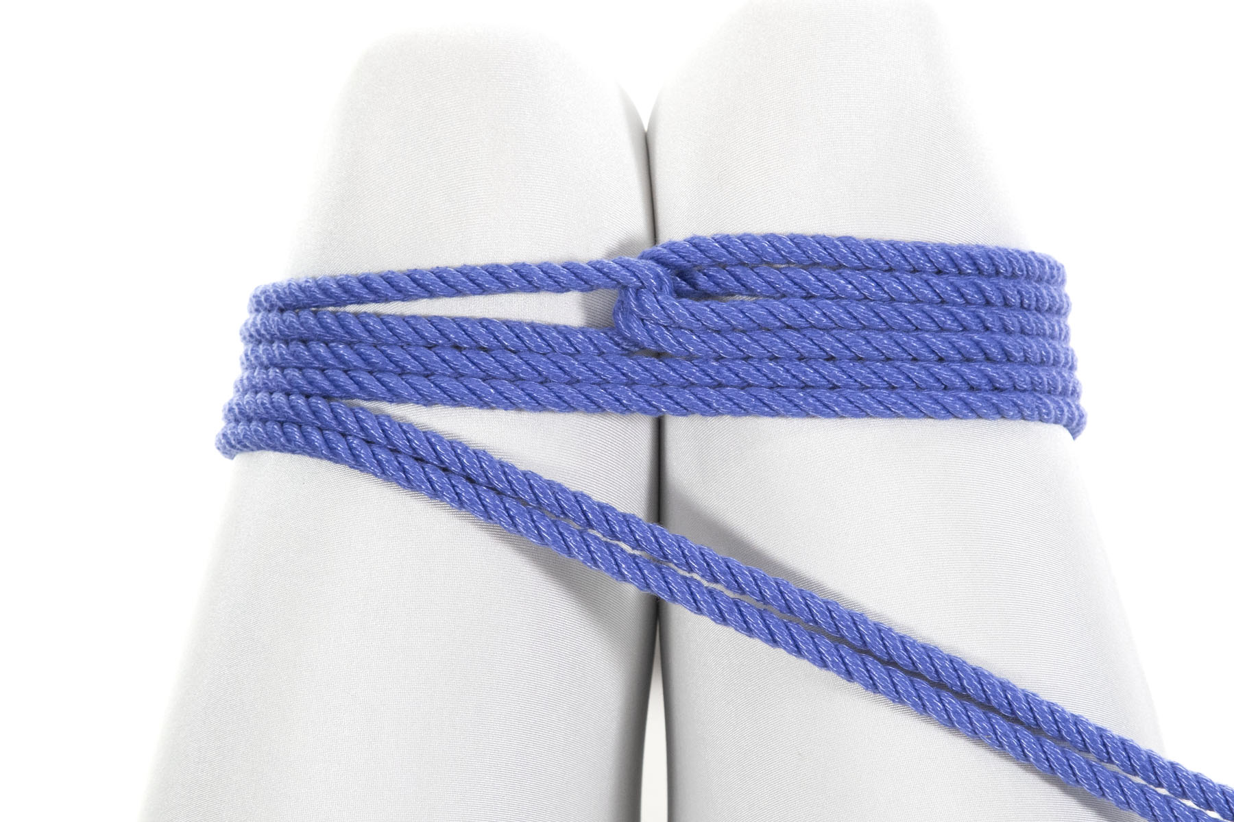 We are looking down at the legs of a person in a white bodysuit, seated in a chair. A doubled blue rope binds the legs together just above the knee. The bight of the rope lies just above the knee and the rope goes around the legs counterclockwise before making a reverse tension and reversing direction. It then makes two complete clockwise spirals, moving toward the body. Each wrap lies flat and adjacent to the previous one. The ends of the rope are pulled out of frame toward the body and to the right.