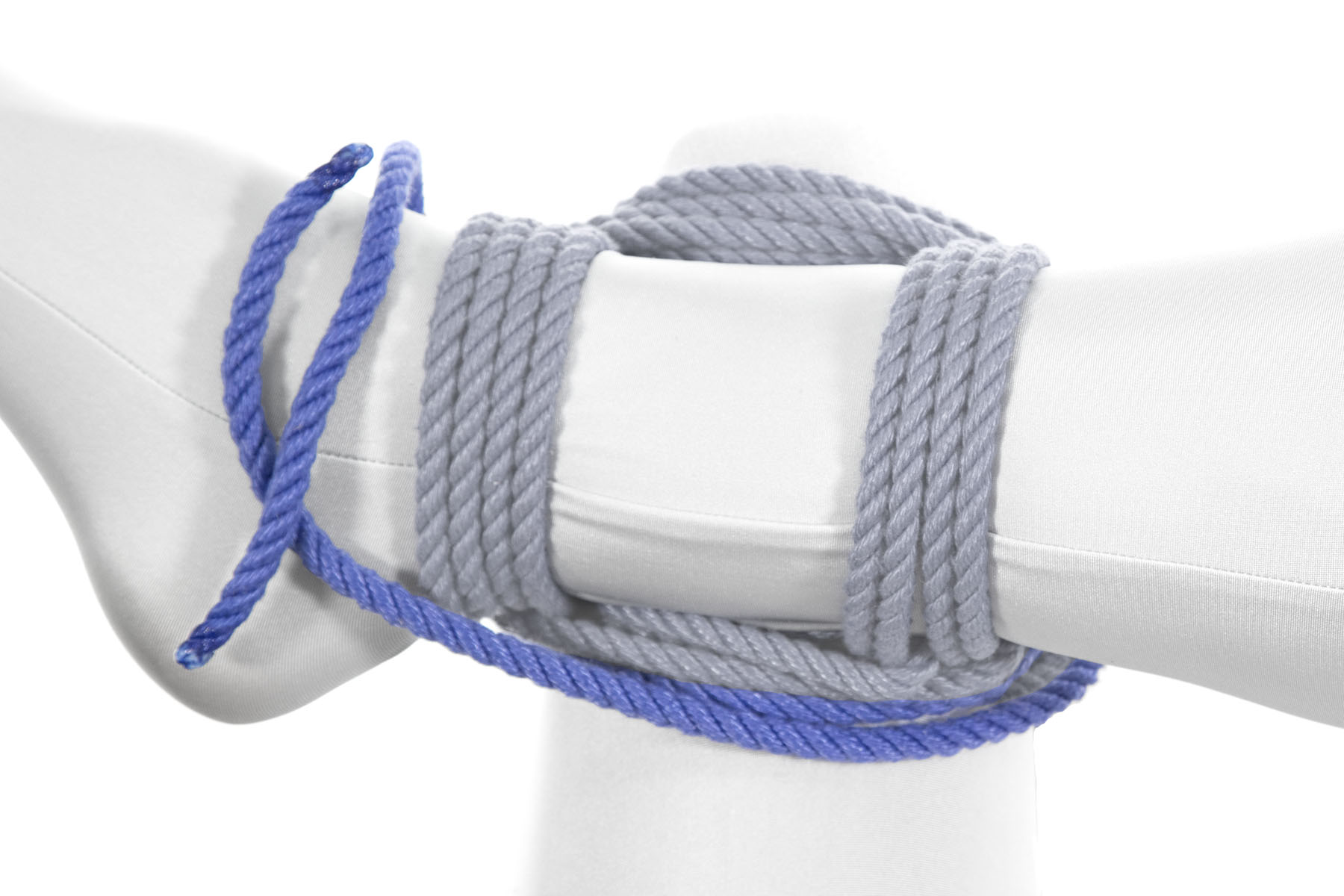 The ends of the rope have been split in preparation for tying a square knot. One end goes diagonally up and over the right ankle, lying on top of it. The other end continues to follow the sqaure, crossing to the left over the left leg and then moving away from the body under the right ankle. It then wraps up around the ankle and meets the first end on top of the ankle.