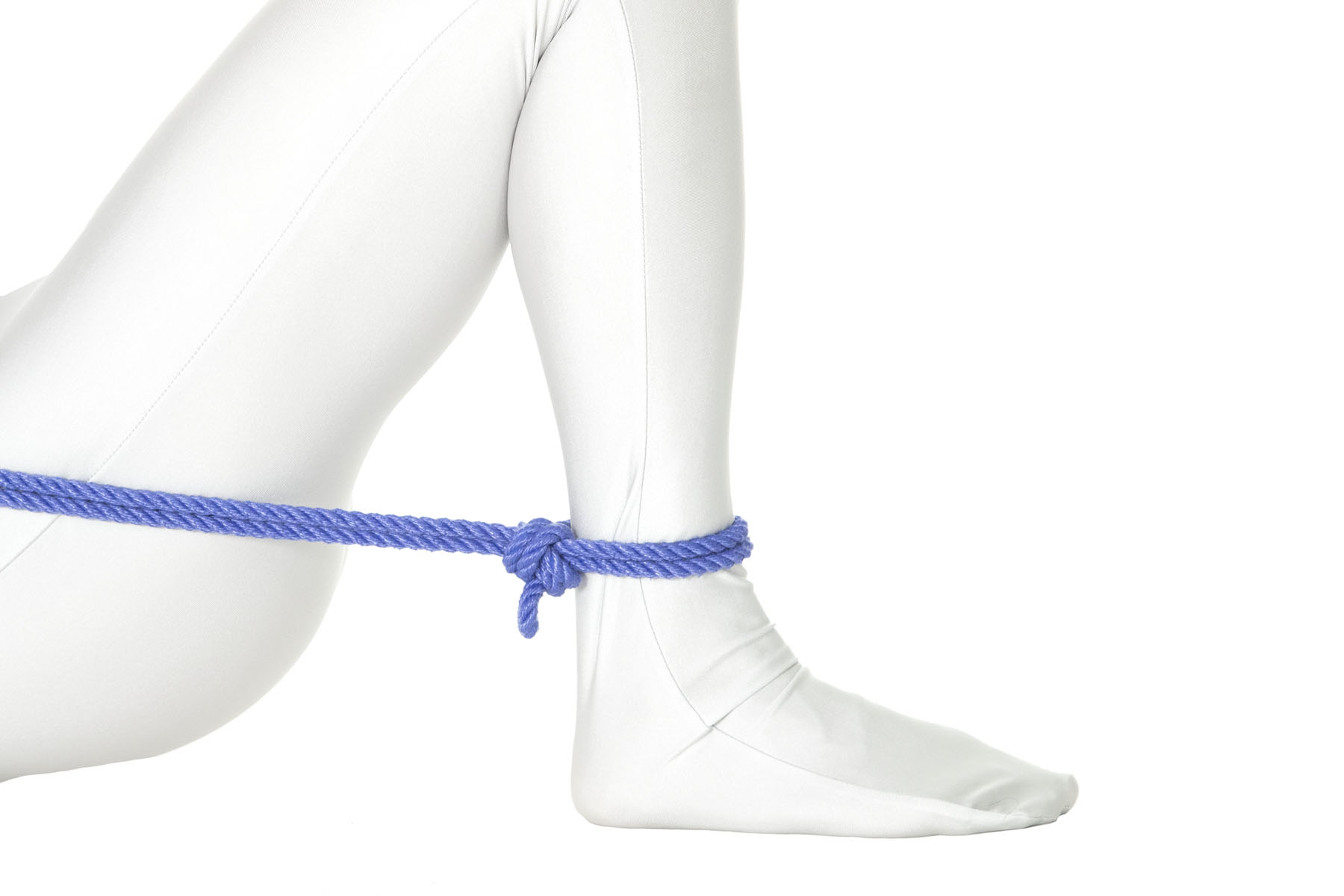 A person in a white bodysuit is lying on their back with their right knee fully bent and their foot flat on the ground. A single column tie is tied around their ankle, with the tail of the rope pulled up toward their body and crossing the thigh just below the hip.
