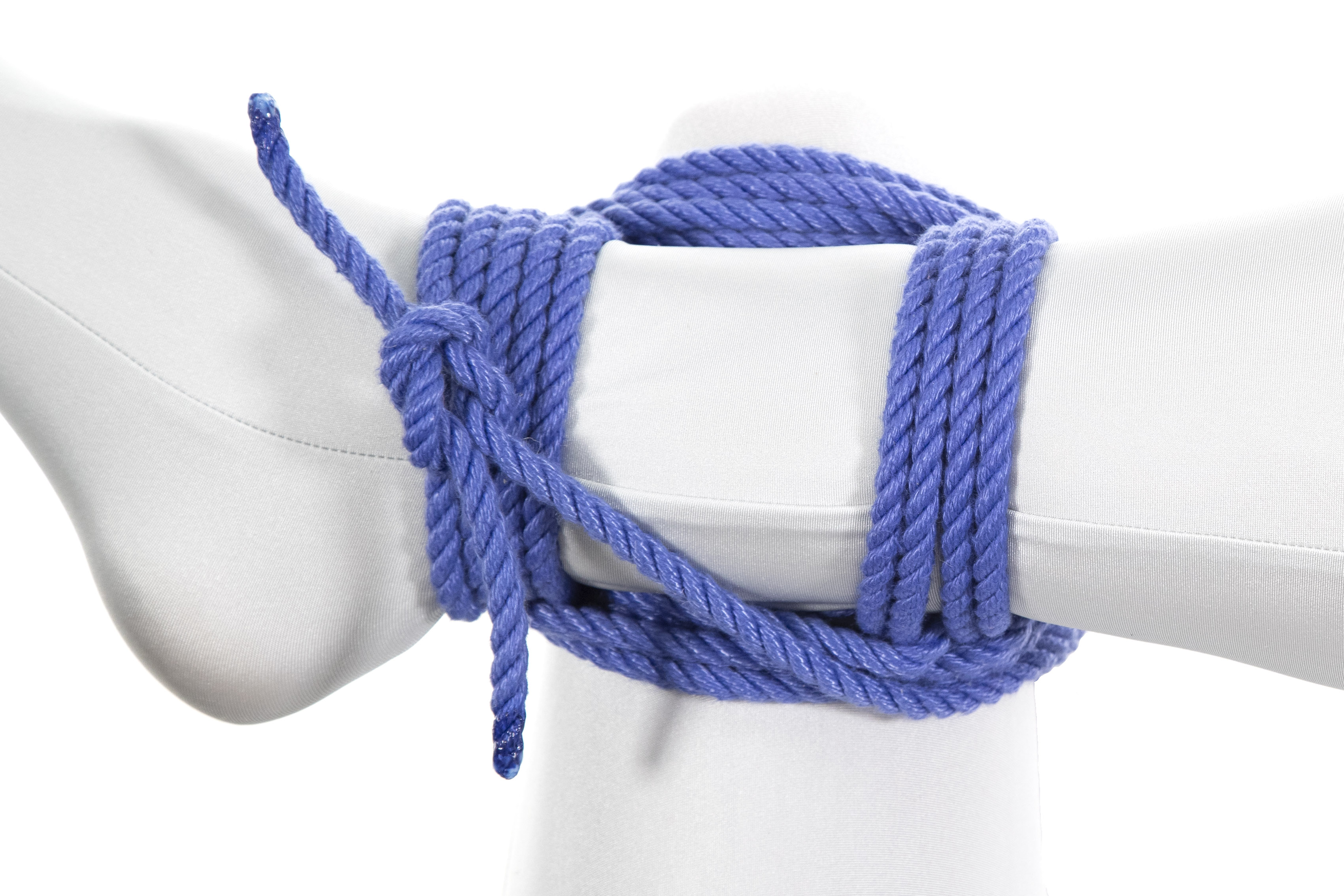 A closeup of an ankle in a white bodysuit tied to a thigh with a square lashing in blue rope.