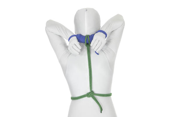 A rear view of a person in a white body suit with their hands tied together behind their head and connected to a rope that runs around their waist.