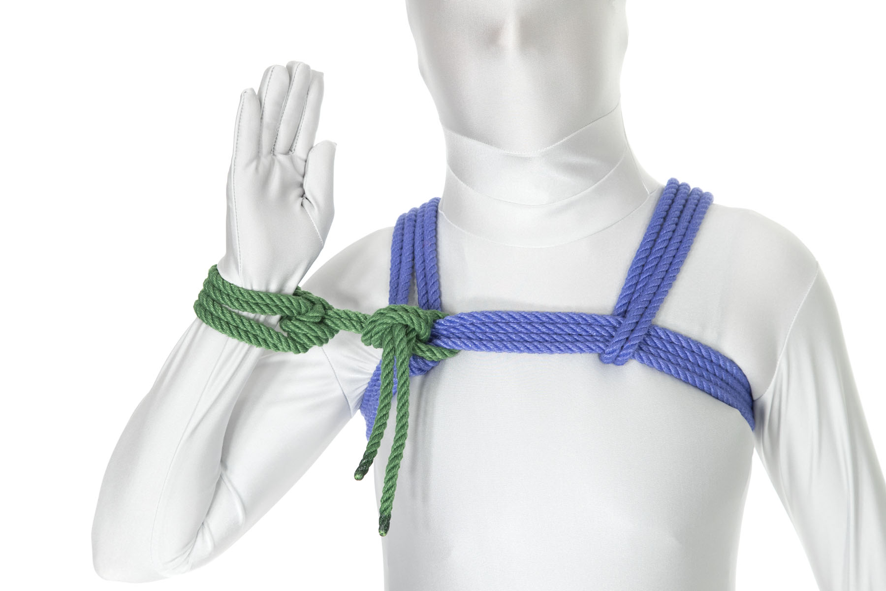 A person in a white bodysuit is raising their right hand with the palm forward, as thought to wave hello. They are wearing a blue chest harness and a green column tie is tied around their right wrist. The rope end from the column tie is tied to the chest harness at the point where the right shoulder line connects to the chest wrap. The column tie is separated from the chest harness by two inches.