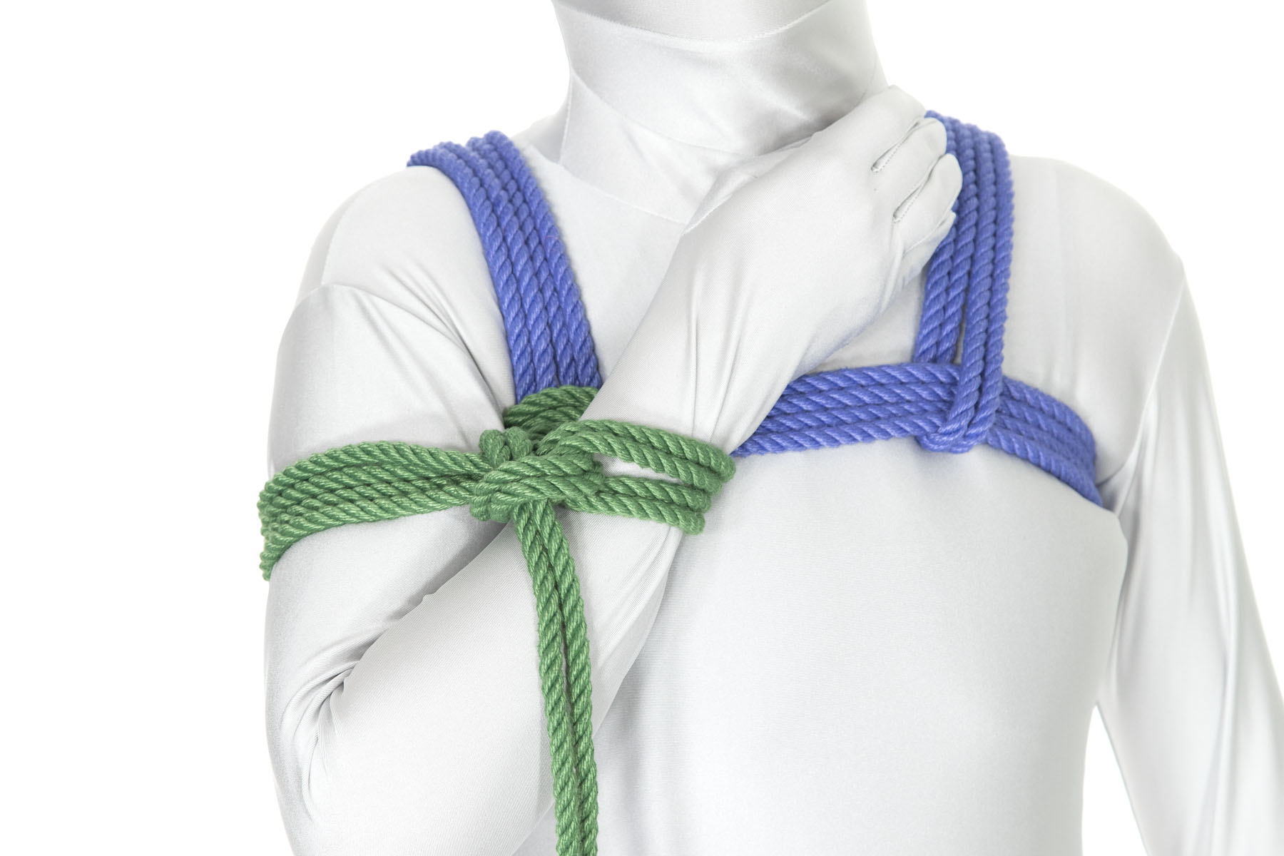 A person in a white bodysuit wearing a blue chest harness. Their right arm is bent tightly at the elbow and is held in front of the body, with the hand over the breastbone. A green column tie binds the upper and lower arm together and is tied off to the chest harness.