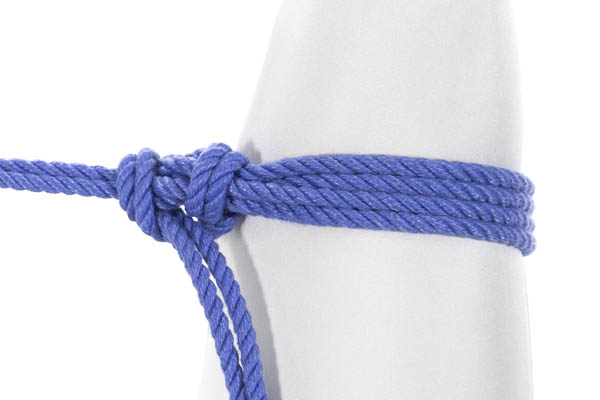 A closeup of a leg in a white bodysuit with a doubled blue rope tied around it in a captured overhand cuff. The rope enters from the left and makes two clockwise wraps around the leg, then makes a complicated knot right where the standing part of the rope meets the wraps. The tail of the rope exits at the bottom of the image.