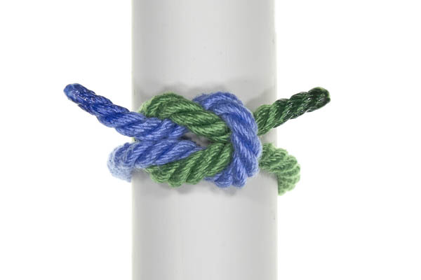 This square knot has been tied around a gray pole that divides the frame vertically. The knot has been pulled tight around the pole and is neat and symmetrical. The blue rope enters from the left, crosses under then over the green rope, makes a loop that crosses twice over the green rope, the crosses under and over the green rope. The working end of the blue rope lies flat and parallel to the standing part, with both crossing over the loop of the green rope. The green rope is a mirror image of the blue rope, but with its loop, working end, and standing part underneath the blue rope. Both standing parts exit on the bottom of the knot and both working ends exit on the top of the knot.