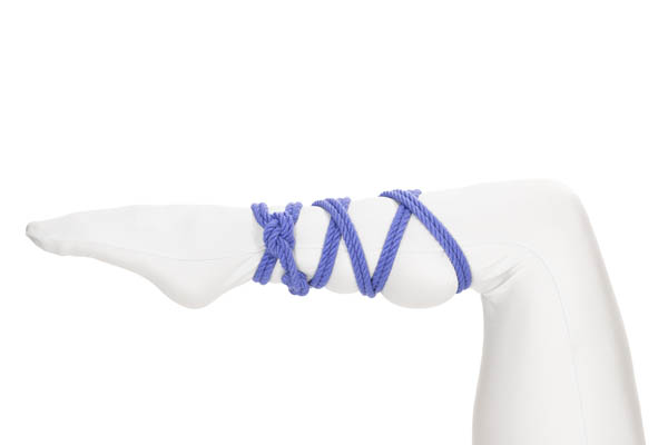 The left leg of a person wearing a white bodysuit enters the image from below and makes a ninety degree bend at the knee, with the lower leg pointing to the left. The foot and toes are pointed. A doubled blue rope is tied around the ankle and spirals up the lower leg and back down again, making X’s where it crosses itself.